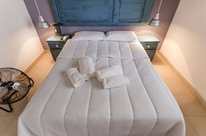 Ein ordentlich bezogenes Bett mit weißer Bettwäsche und aufgerollten Handtüchern, ein Nachttisch mit einer Lampe und ein Ventilator auf dem Boden.