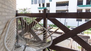 Un balcon avec une chaise en fil de fer blanc, une balustrade en bois et une vue sur un immeuble doté de nombreuses fenêtres.