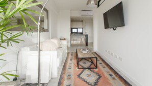 Un salon moderne avec un canapé blanc, une table basse et un grand lampadaire. On aperçoit à l’arrière-plan un coin cuisine avec une table à manger et des chaises.