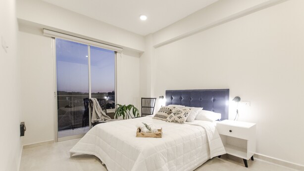 Ein Schlafzimmer mit einem großen Bett, einem Nachttisch, einer Schiebetür zum Balkon und Blick auf die Stadt im Abendlicht.
