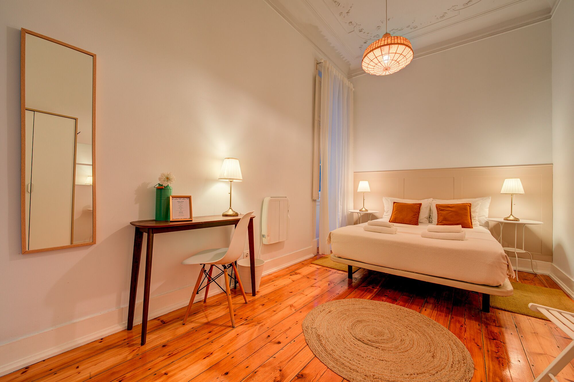 A bedroom with a large bed, two bedside tables with lamps, a wooden floor, a round rug, and a mirror on the wall.
