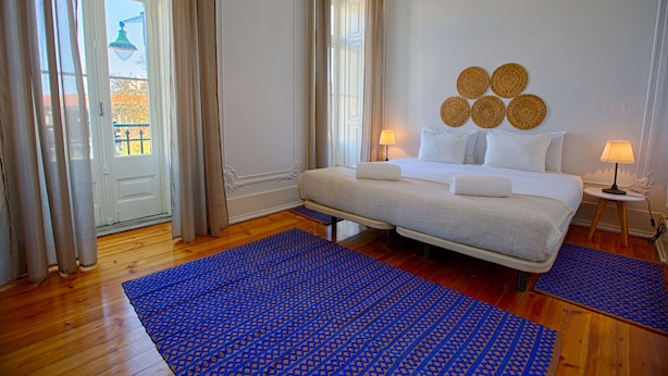 Quarto de hotel com uma cama, duas luminárias de cabeceira, um tapete azul estampado e vista para o exterior através de uma janela.
