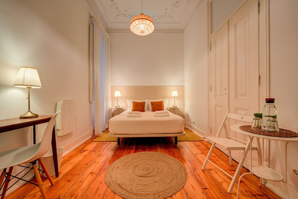 A hotel room with a bed, bedside tables, a desk, a chair, a round rug, and a hanging light fixture.
