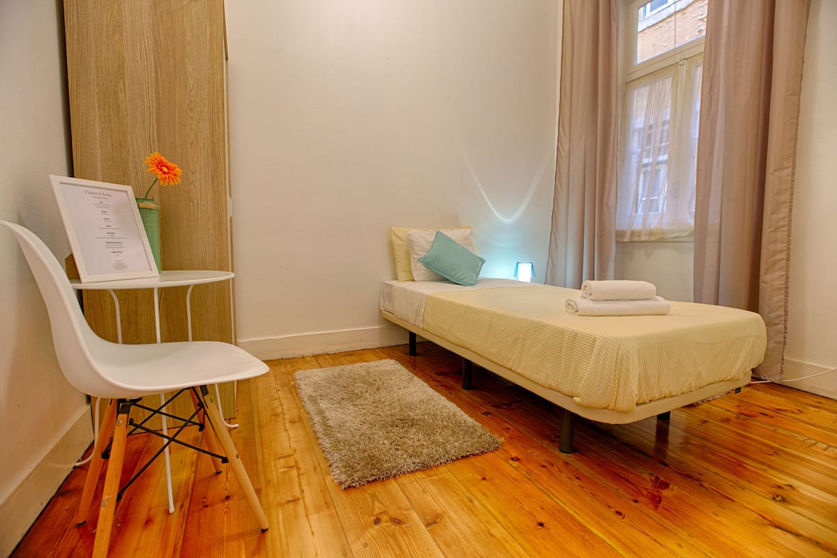 A small, neatly arranged bedroom with a single bed, a chair, a small table with a menu, and a window with curtains.