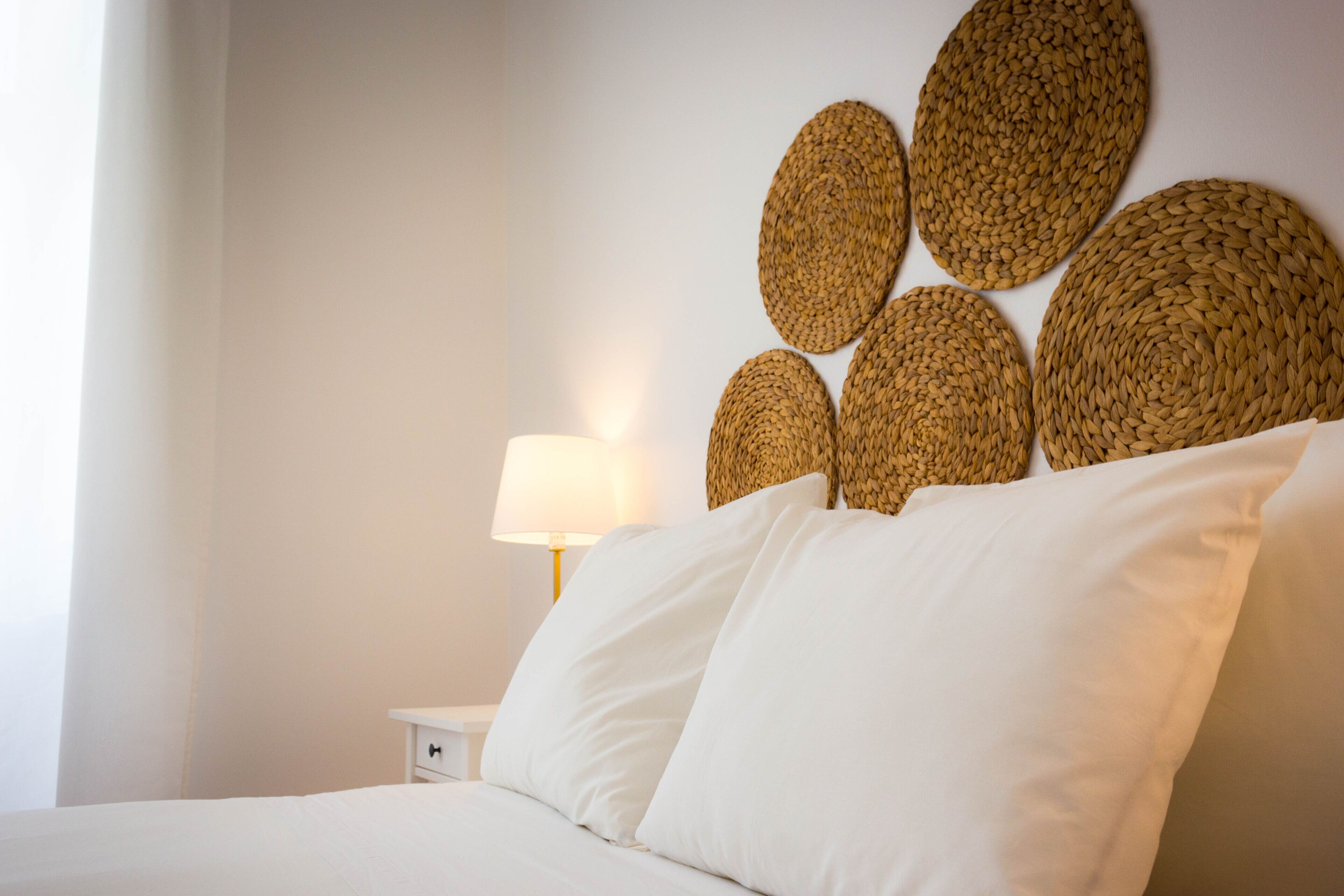 Un lit bien fait, avec du linge de lit blanc, une table de chevet avec une lampe et un mur décoré de motifs circulaires tissés.