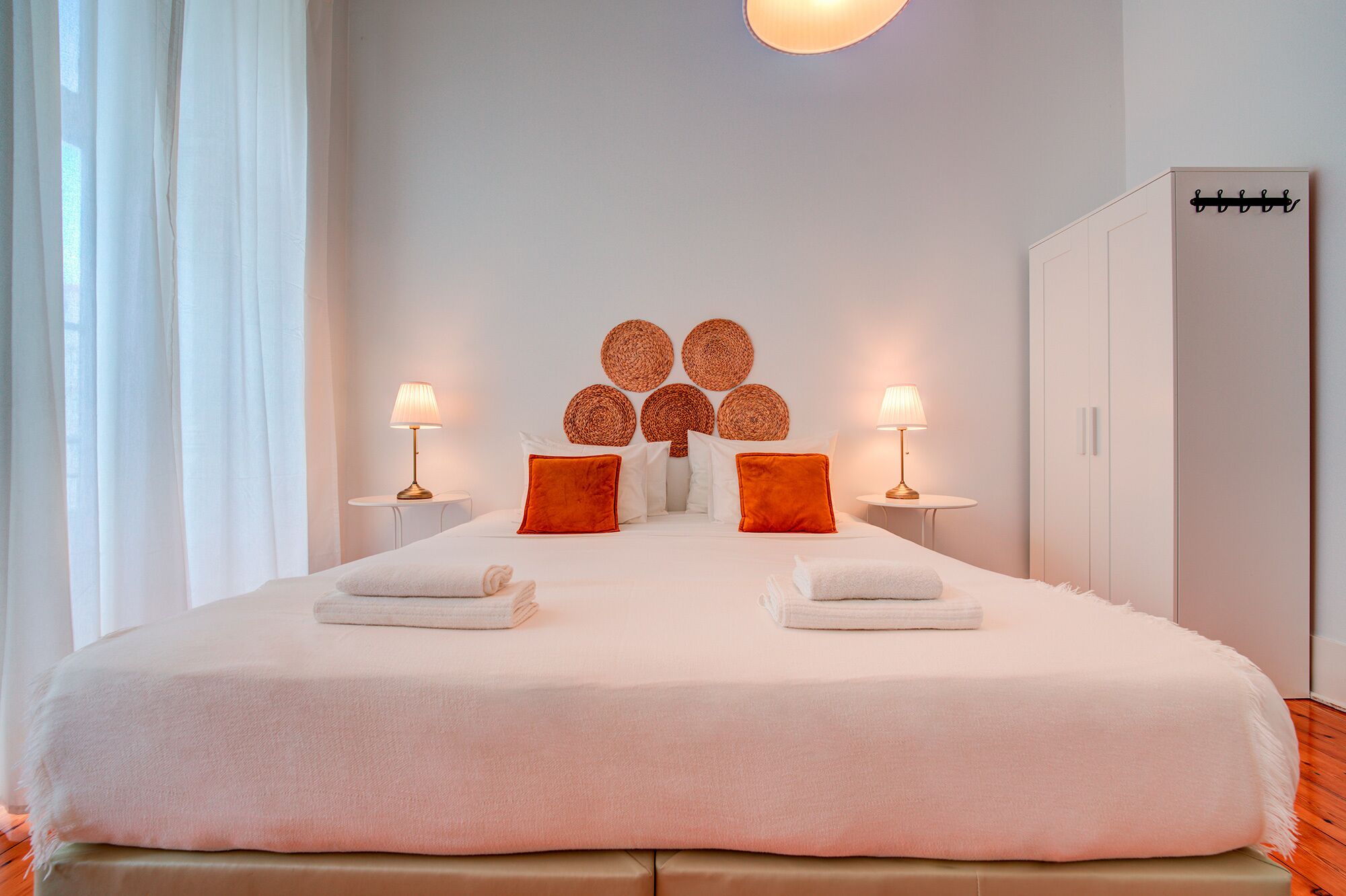 Una cama bien tendida con sábanas blancas y dos toallas, una mesita de noche con lámpara, y una pared decorada con arte de pared tejido.