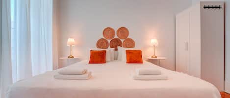 Una cama bien tendida con sábanas blancas y dos toallas, una mesita de noche con lámpara, y una pared decorada con arte de pared tejido.