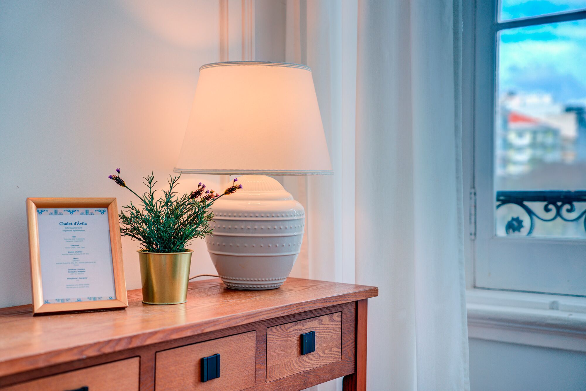 Une table de chevet en bois, sur laquelle on trouve un menu encadré, une plante en pot et une lampe blanche.