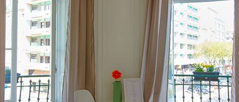 Habitación de hotel con cama, escritorio y silla, un florero con una flor, y vista de la ciudad a través de amplias ventanas.