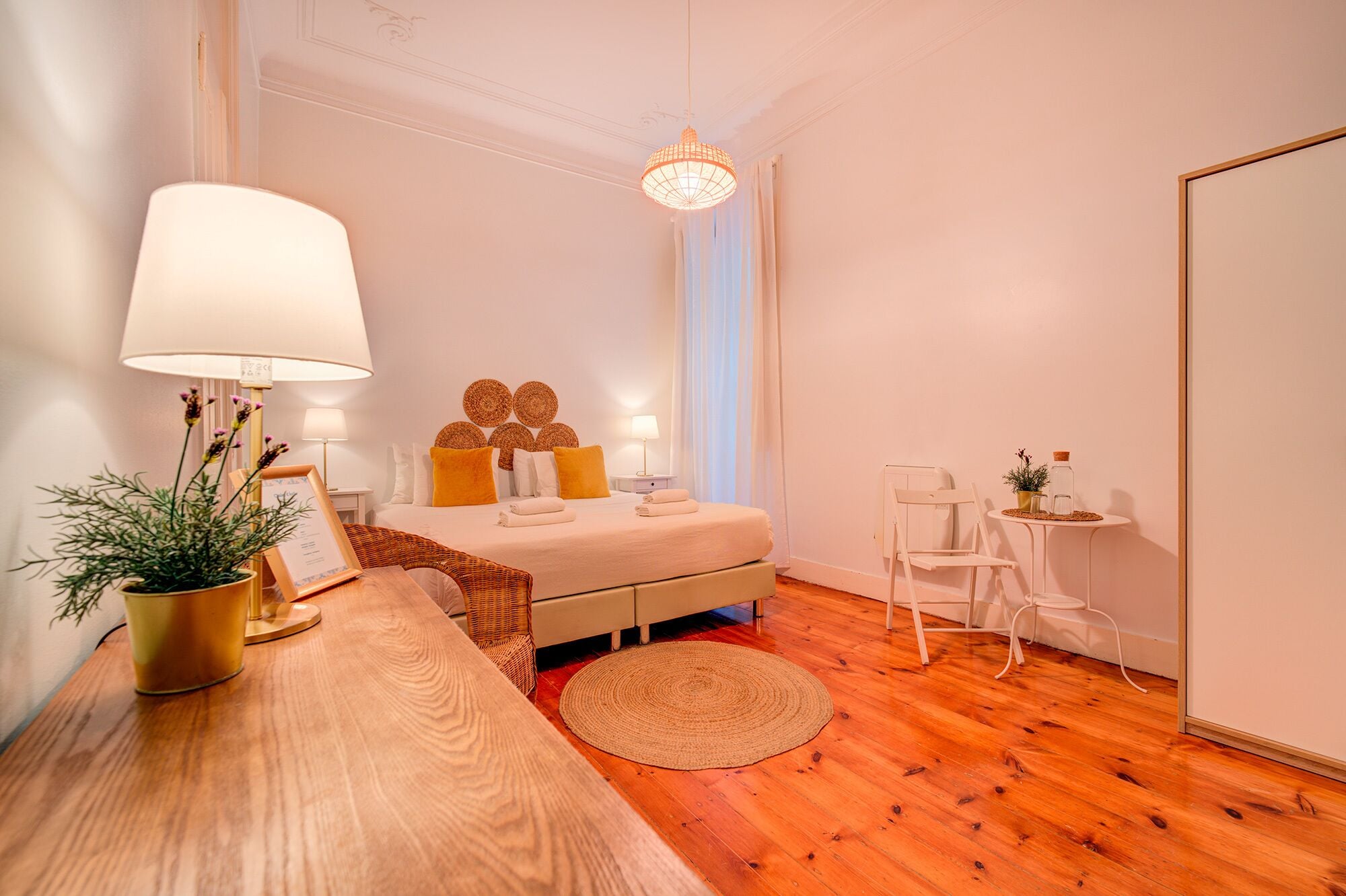 Un dormitorio bien iluminado con piso de madera, una cama con ropa de cama blanca y amarilla, una mesita de noche con una lámpara, una pequeña mesa con una planta y una silla.