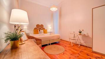 Un dormitorio bien iluminado con piso de madera, una cama con ropa de cama blanca y amarilla, una mesita de noche con una lámpara, una pequeña mesa con una planta y una silla.