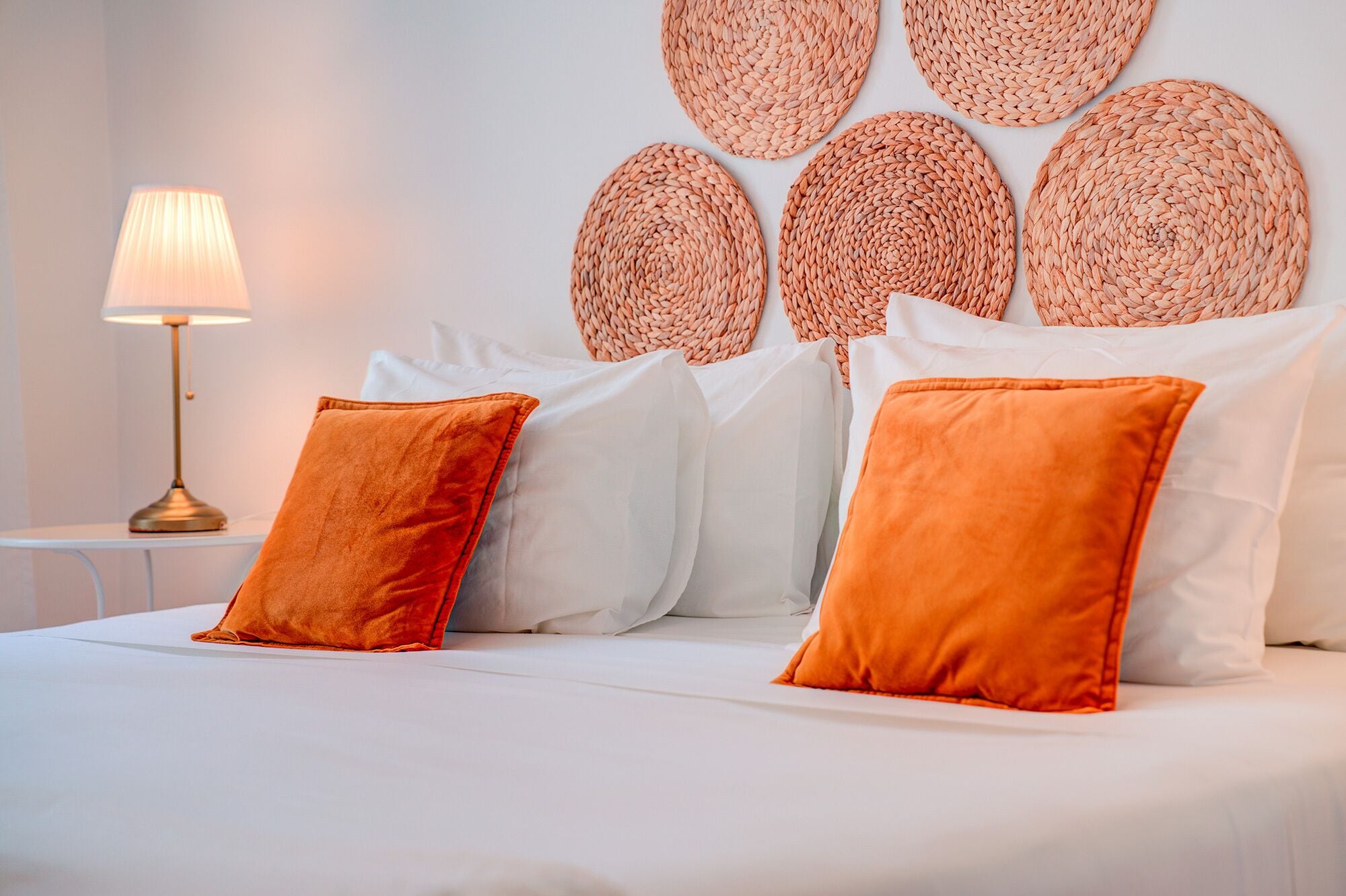 Una cama bien tendida con almohadas blancas y anaranjadas, una lámpara de noche y un panel decorativo tejido en la pared.
