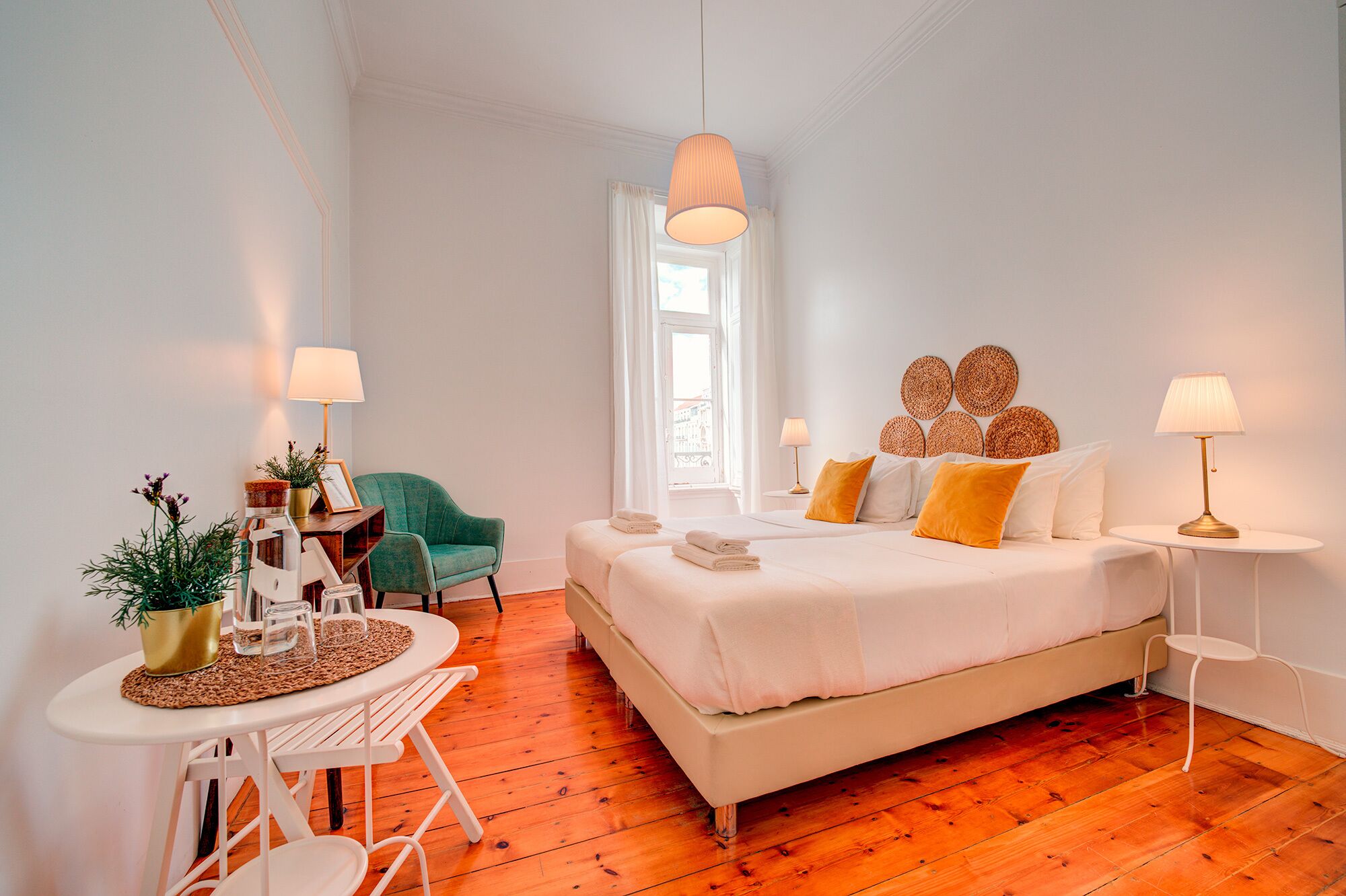 A modern bedroom with a large bed, bedside lamps, a green armchair, a small round table with a plant, and a view of the city through a window.