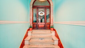 Escada que leva a uma porta de madeira com painel de vidro, ladeada por paredes azul-claras e uma luminária decorativa no teto.