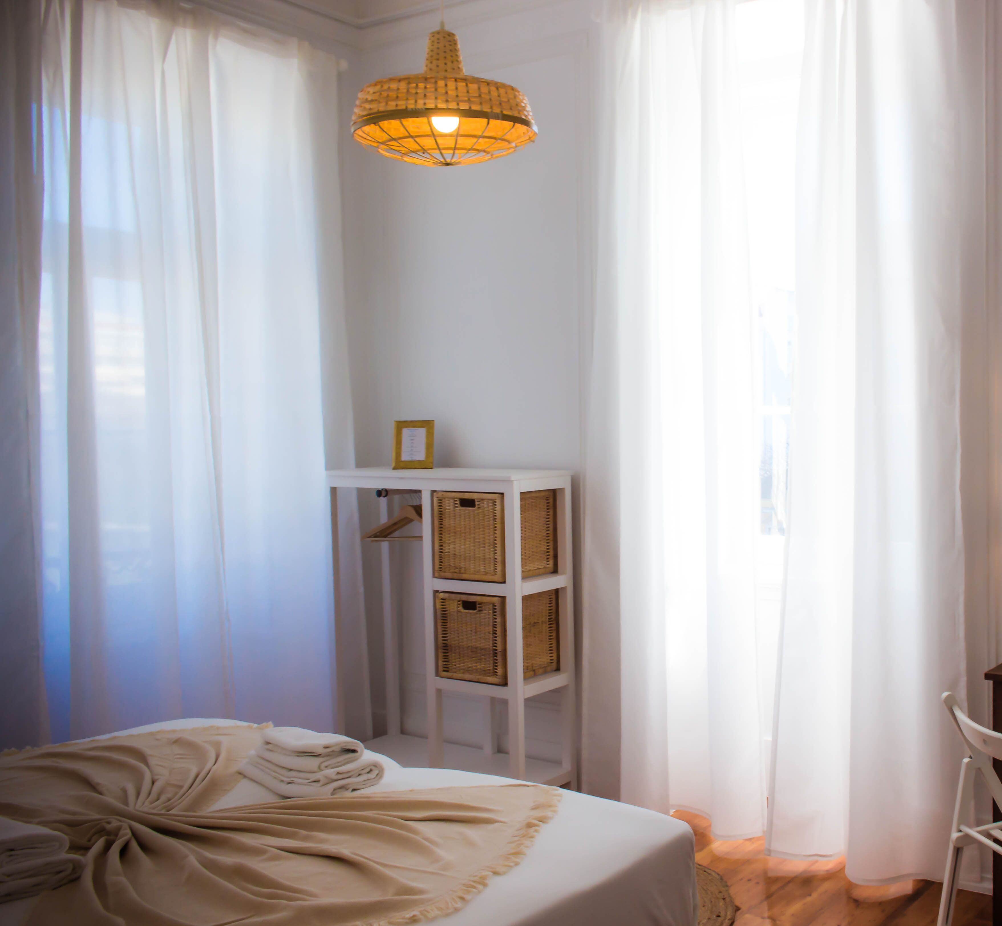 Une chambre à coucher avec un lit, un luminaire suspendu, une étagère blanche avec des paniers et des rideaux légers.