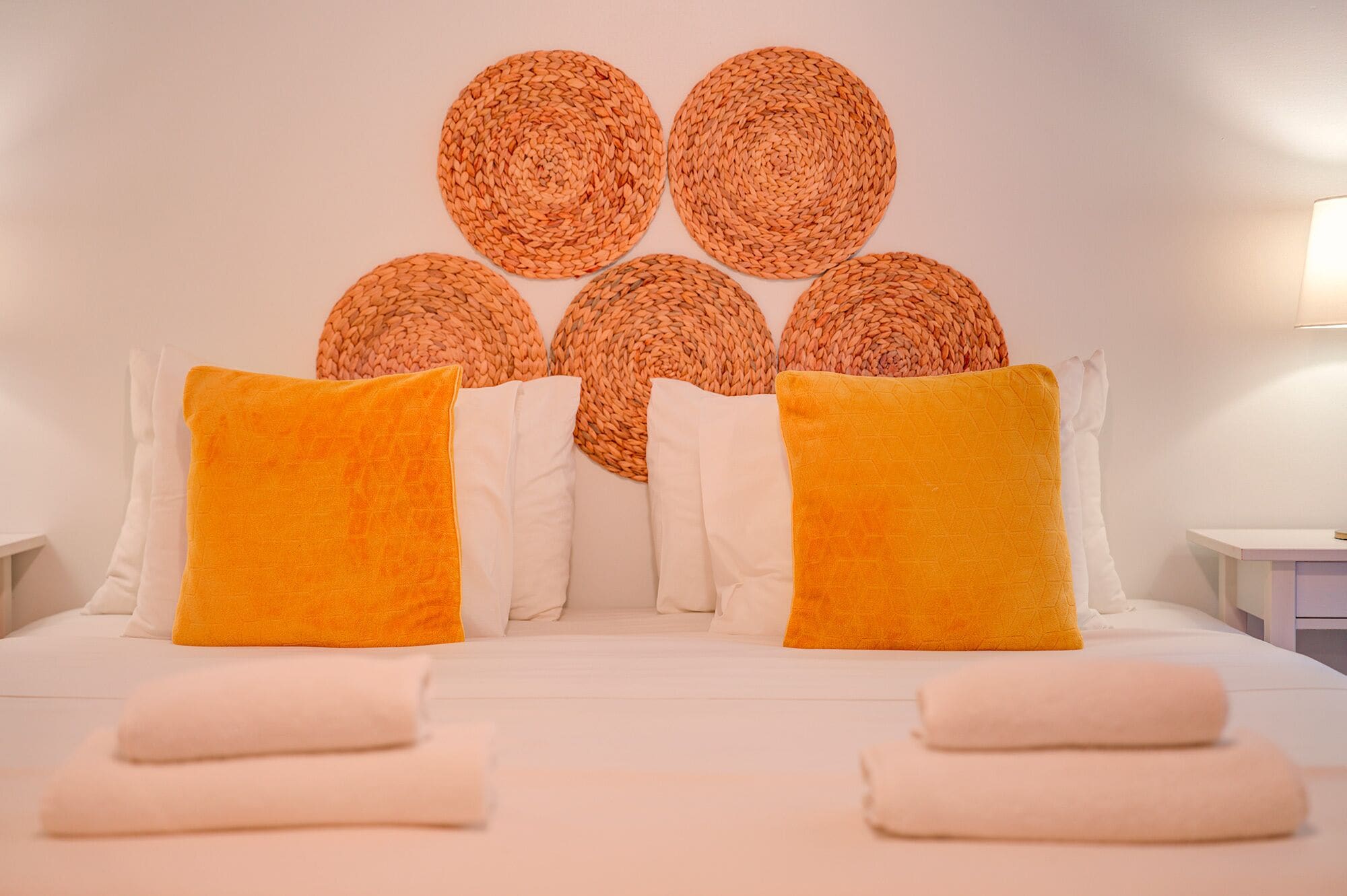 Una cama con sábanas blancas, dos almohadas naranjas y un cabecero tejido.