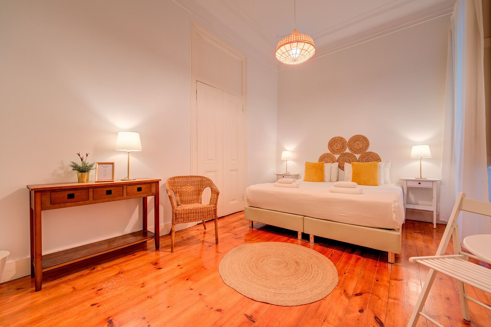 A bedroom with a wooden floor, a bed with white bedding, a bedside table with a lamp, a chair, and a hanging light fixture.