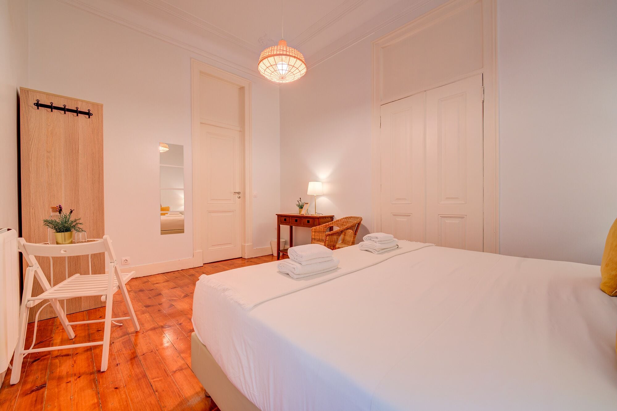 A bedroom with a large bed, a wooden wardrobe, a small table with a lamp, a chair, and a mirror on the wall.