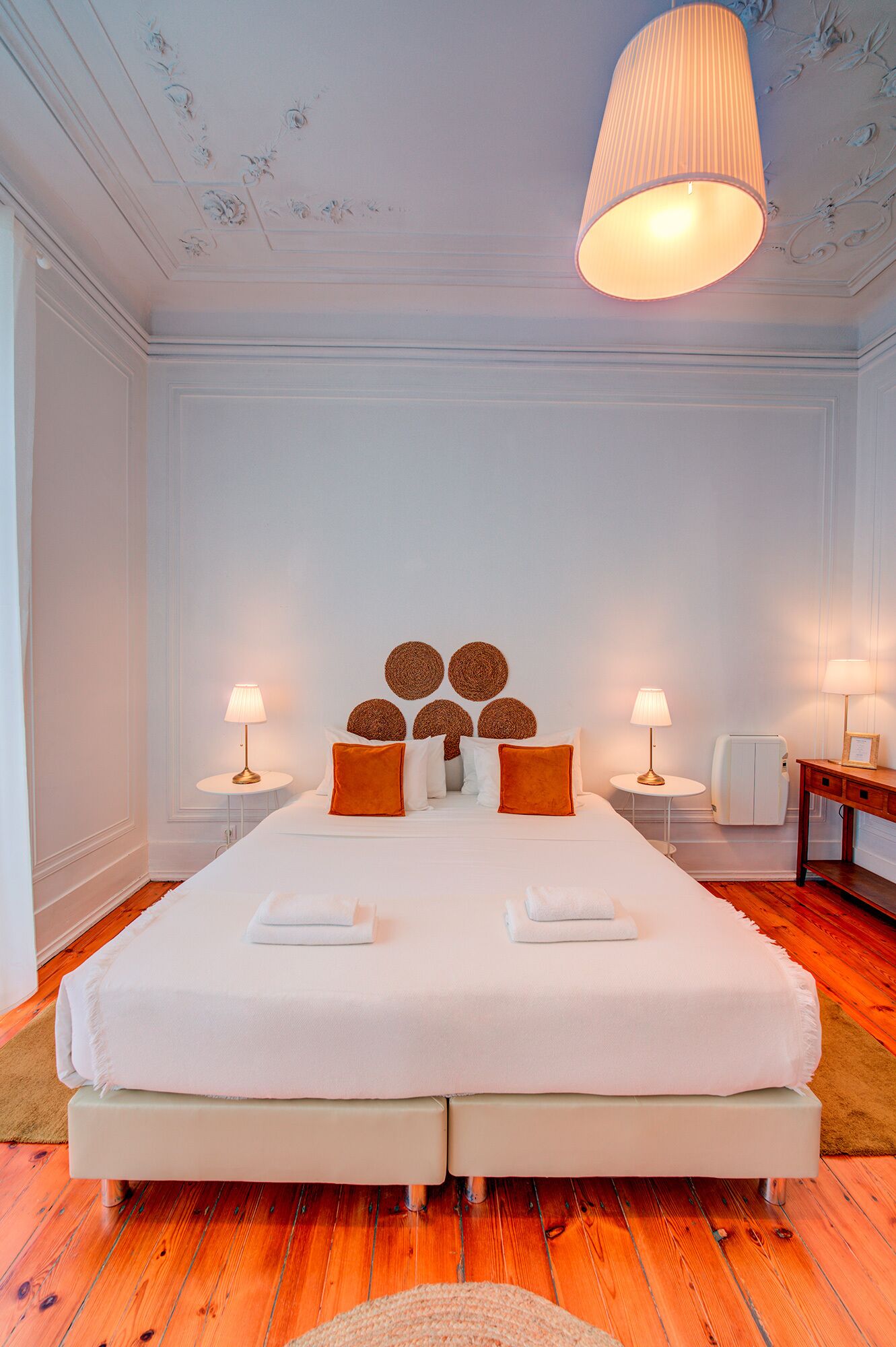A double bed with white linens, two bedside tables with lamps, a round wall decoration, and a ceiling light fixture.