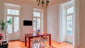 Um quarto amplo com piso de madeira, uma grande mesa de jantar com cadeiras, um lustre e janelas amplas que proporcionam luz natural.