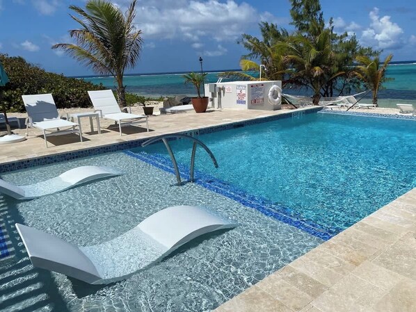 A pool with clear blue water, surrounded by white lounge chairs and palm trees, with a view of the ocean.