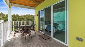 Un balcón cubierto con una mesa y sillas, una puerta corrediza de vidrio que da a un dormitorio y vistas a zonas verdes.