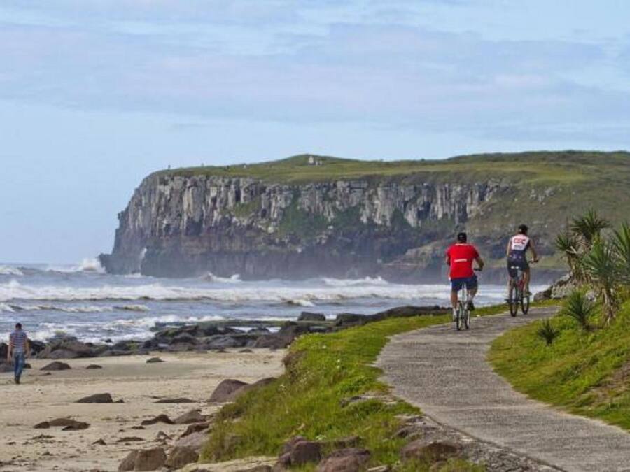 Camino costero con ciclistas y una persona caminando, que conduce a un acantilado con vistas al océano.