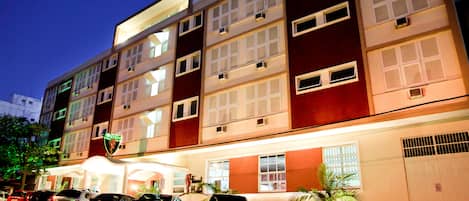 A multi-story hotel with a modern facade, multiple balconies, and a parking area with several cars.