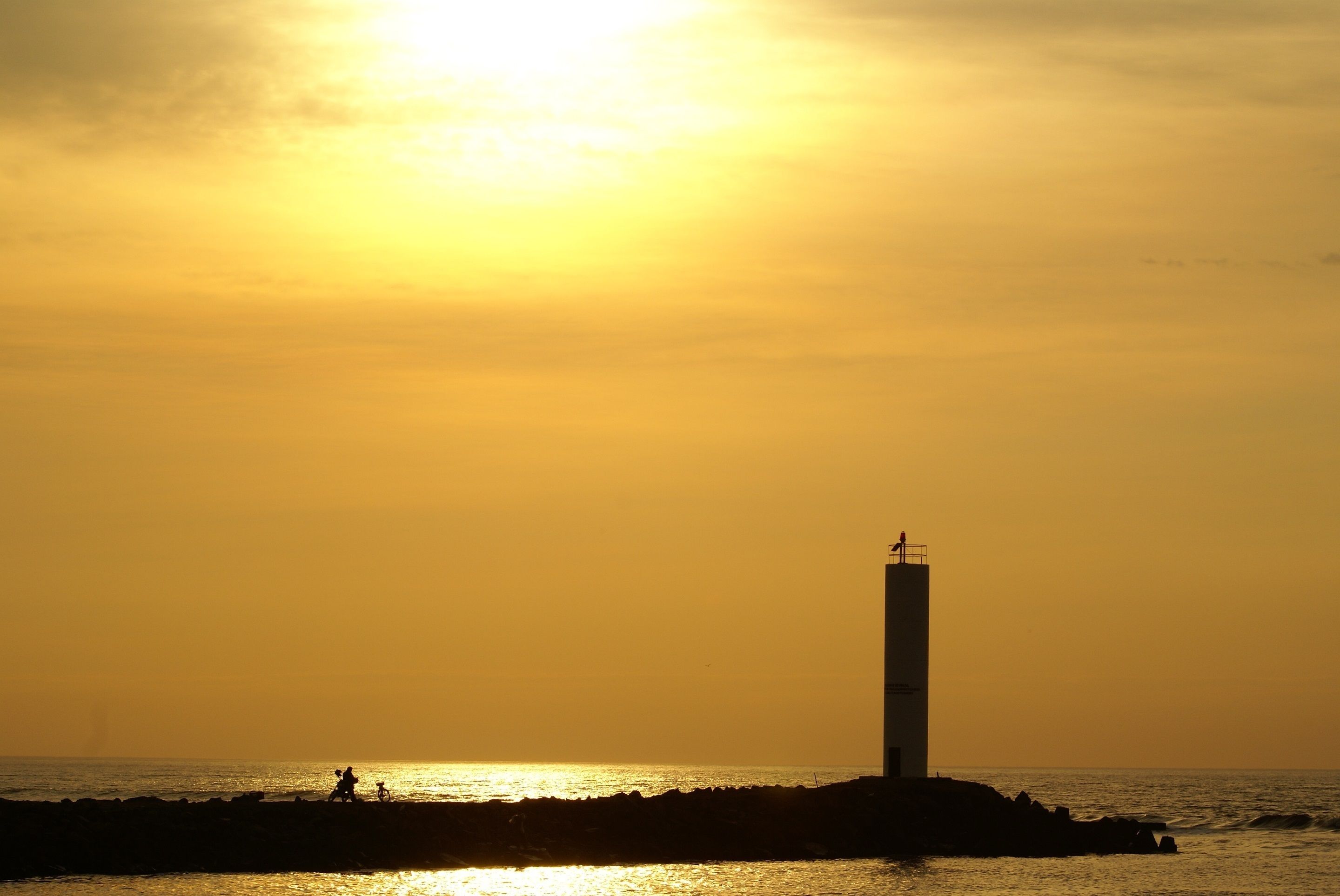 A lighthouse silhouette against a sunset, with a rocky shoreline and a few people in the distance.