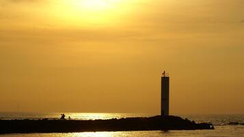 Silueta de un faro contra un atardecer, con una costa rocosa y algunas personas a lo lejos.