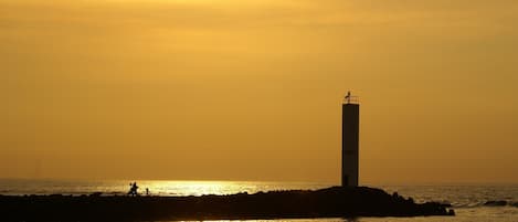 A lighthouse silhouette against a sunset, with a rocky shoreline and a few people in the distance.