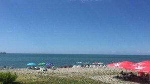 Une plage bordée de rangées de transats et de parasols, un navire à l’horizon et un ciel bleu dégagé.