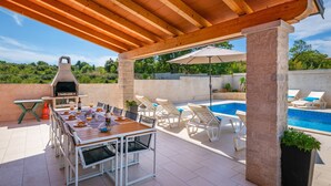 An outdoor patio with a wooden pergola, a barbecue grill, a dining table with chairs, and a pool area with lounge chairs. - Family villa Nicole in Štinjan (Štinjan)