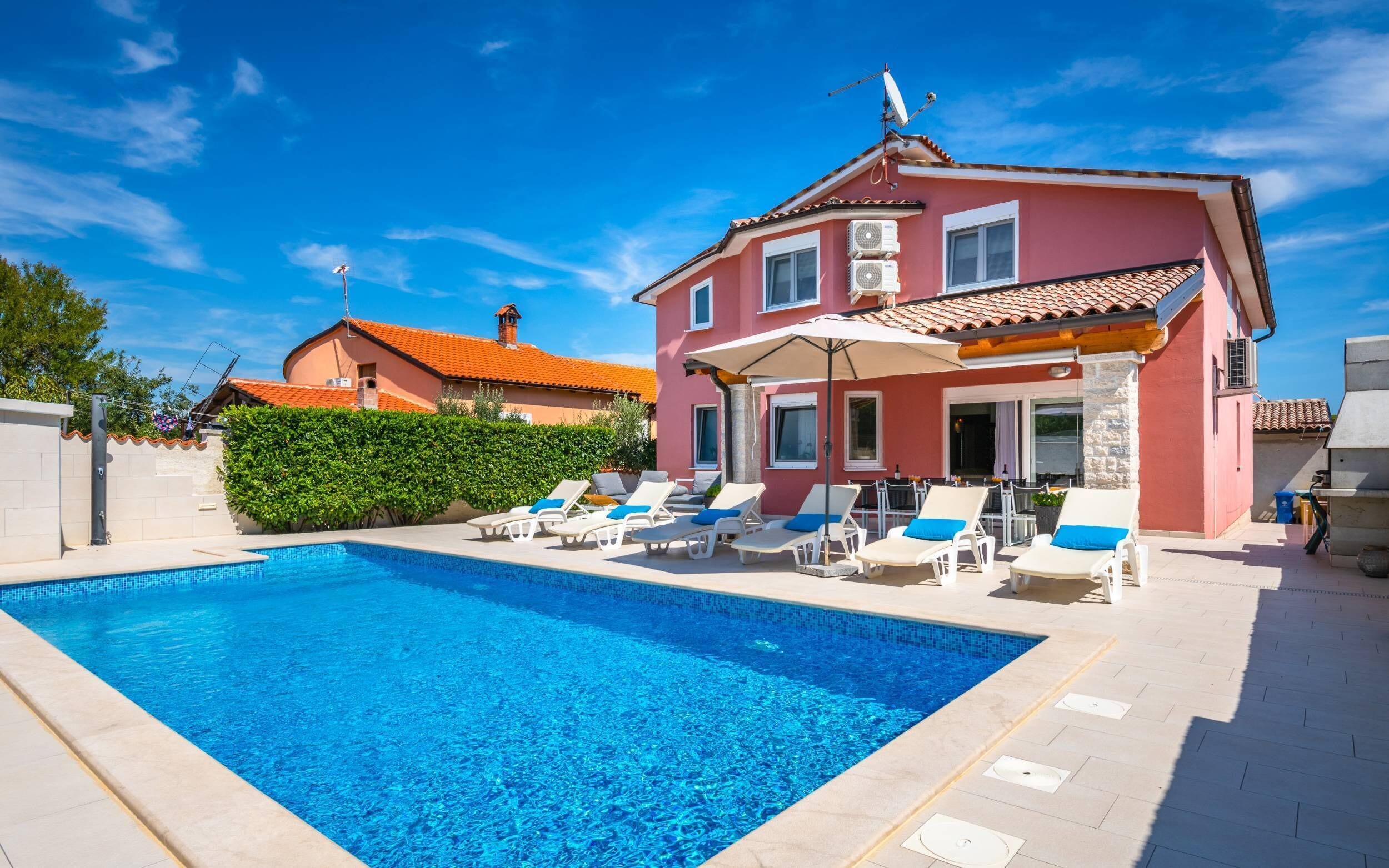 A swimming pool with lounge chairs and umbrellas, a pink building with a tiled roof, and a clear blue sky.