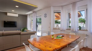 A modern living room with a wooden dining table, white chairs, a beige sofa, a flat-screen TV, and large windows with curtains. - Family villa Nicole in Štinjan (Štinjan)