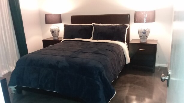 Quarto com edredom azul escuro, duas mesas de cabeceira com abajures e uma mesinha de apoio com um vaso decorativo.