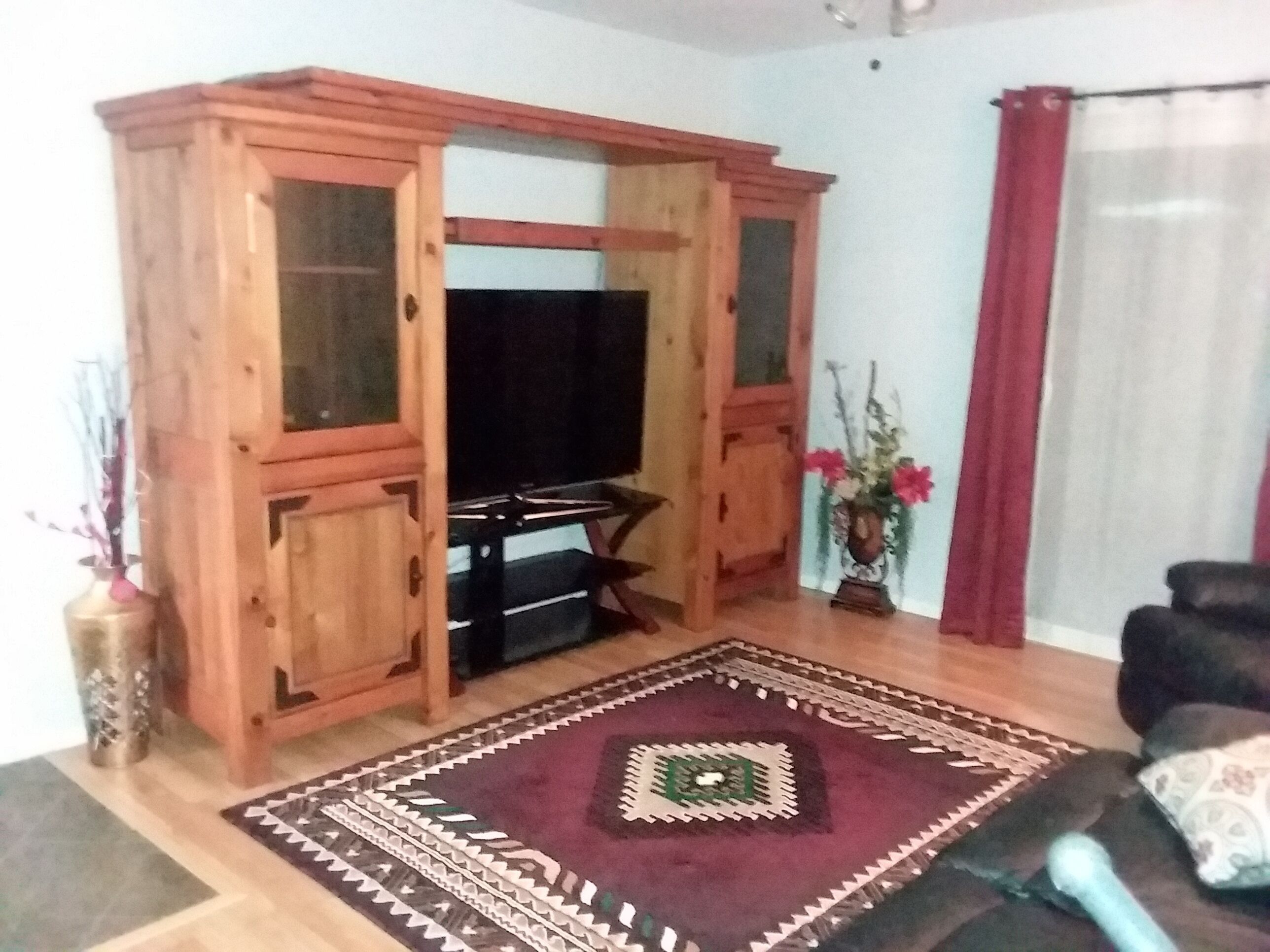A living room with a wooden cabinet, a flat-screen TV, a patterned rug, and a red curtain.