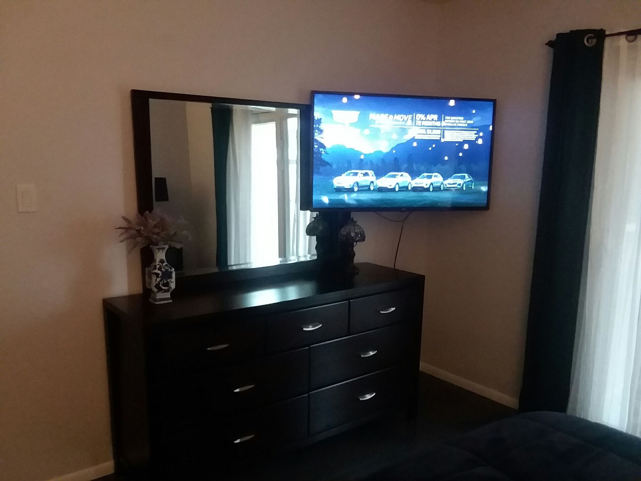 A bedroom with a TV mounted on the wall, a dresser with drawers, and a vase with flowers on the dresser.