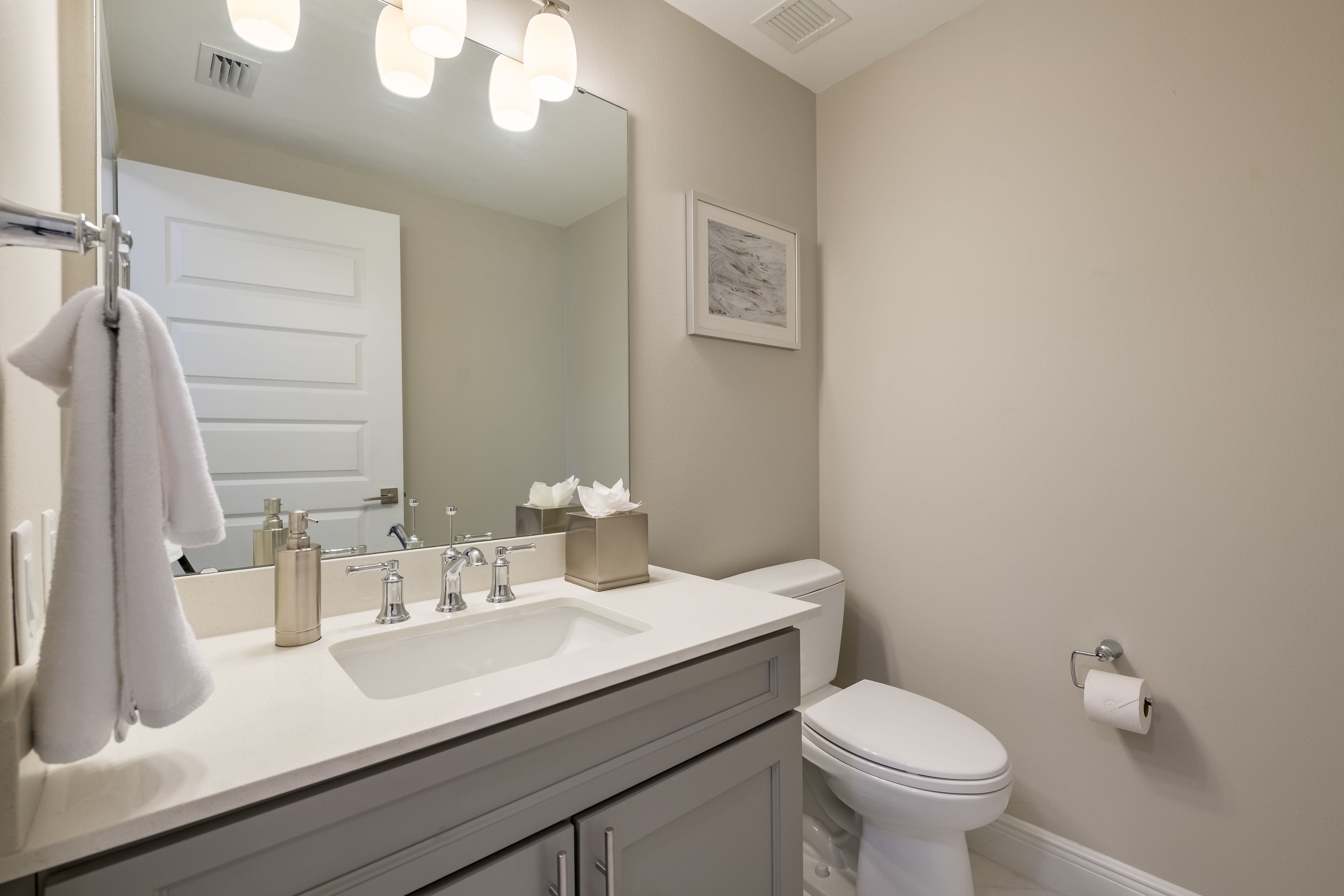 A bathroom with a white sink, mirror, and toilet. There is a towel hanging on the wall and a framed picture above the toilet.