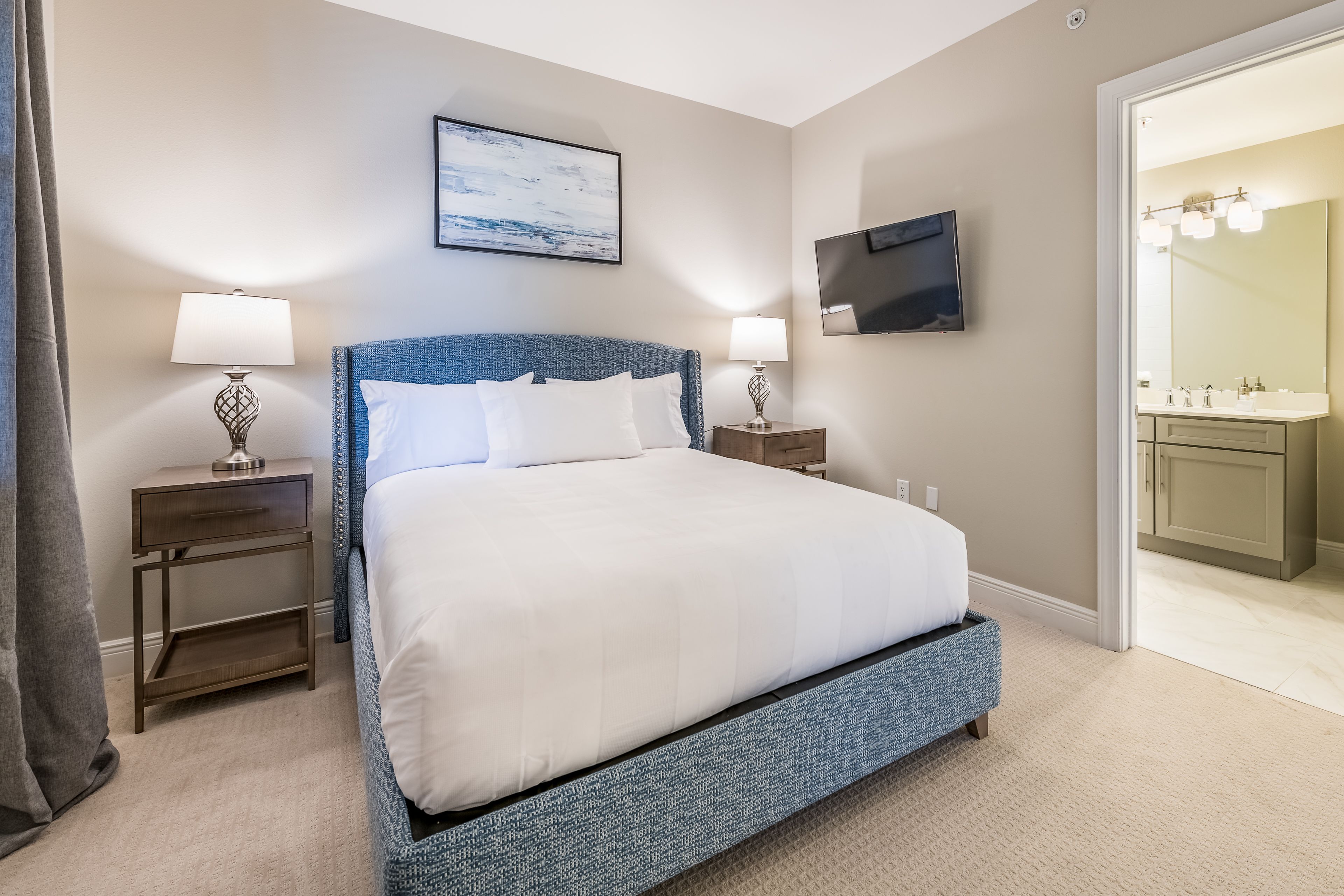 A bedroom with a bed, two bedside tables, a TV, and a bathroom visible through an open door.