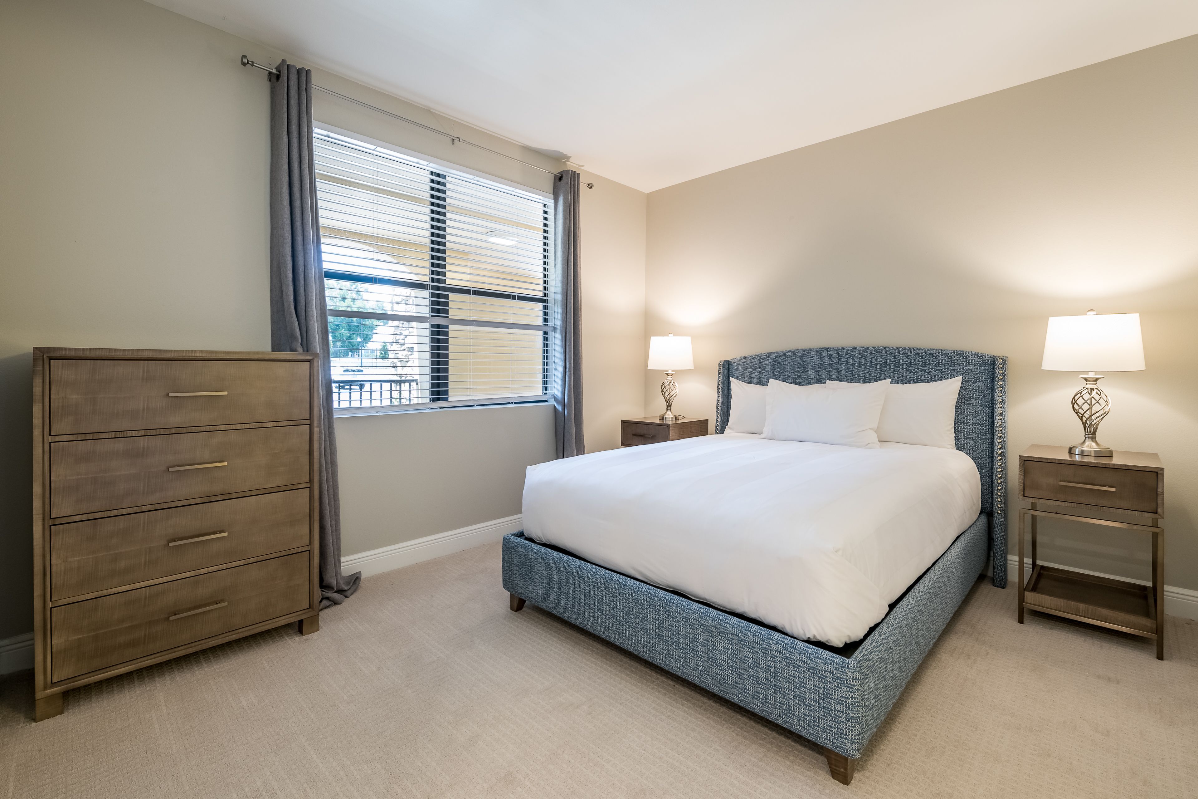 A bedroom with a large bed, a dresser, two bedside tables with lamps, and a window with blinds.