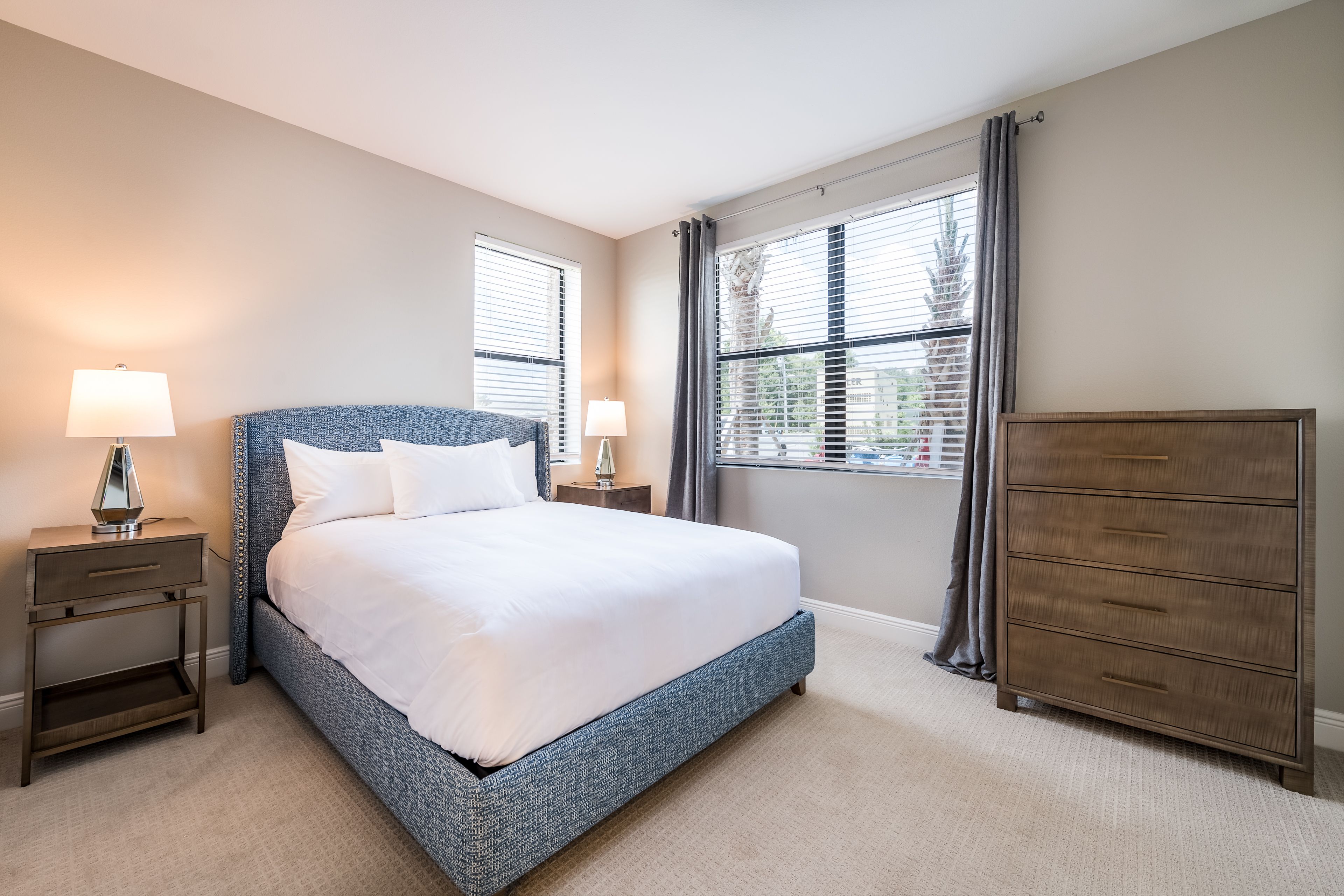 A bedroom with a large bed, two bedside tables with lamps, a dresser, and a window with blinds.
