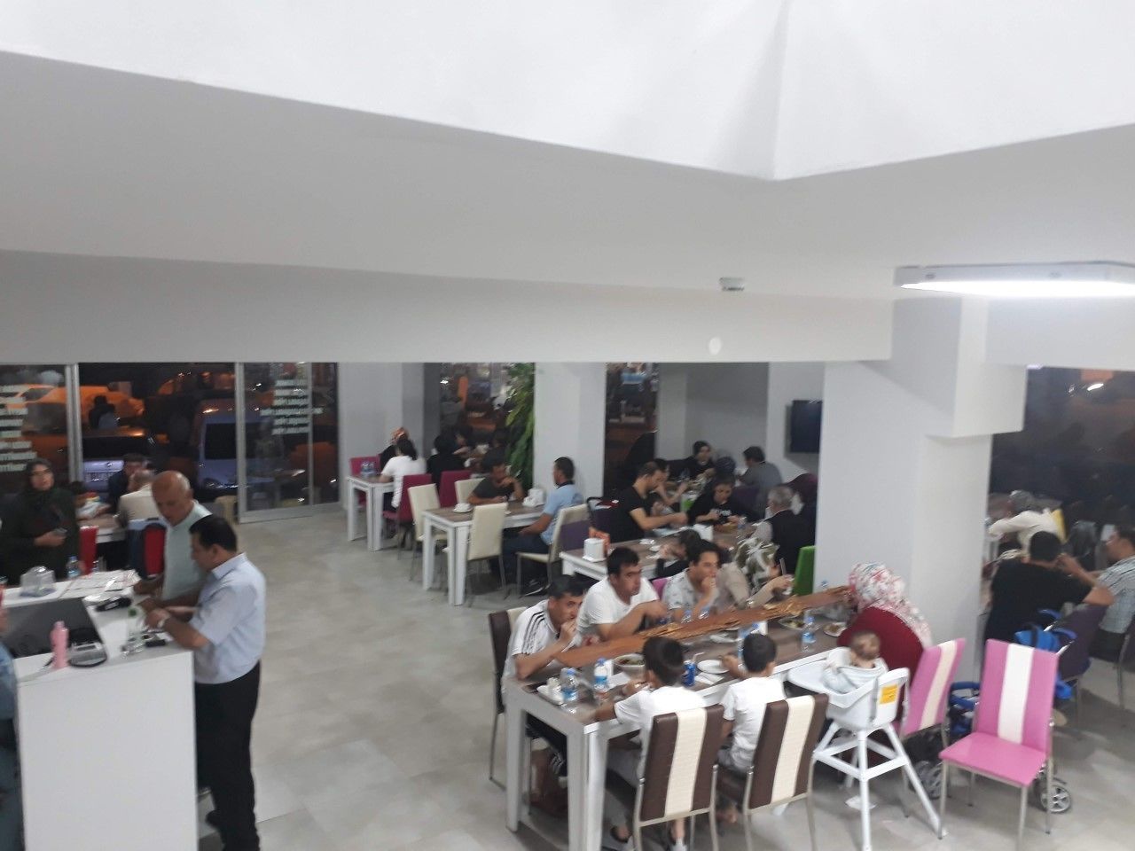 Un vaste espace dédié à la salle à manger, avec de nombreuses tables et chaises, un comptoir où se trouve un membre du personnel, et de grandes fenêtres qui laissent entrer la lumière naturelle.