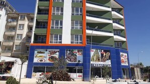 Een meerlaags gebouw met balkons, een restaurant genaamd "KEBAPISTAN" op de begane grond, en een helderblauwe lucht op de achtergrond.