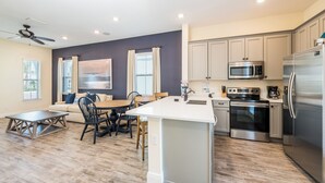 A modern kitchen with a central island, stainless steel appliances, and a dining area with a wooden table and chairs. - Lovely Cottage near Disney | Hot Tub + Resort & Waterpark Access - 8092DR (Kissimmee)