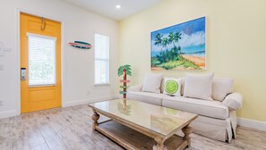 A living room with a glass coffee table, a beige sofa with cushions, a painting of a beach scene, and a door with blinds.