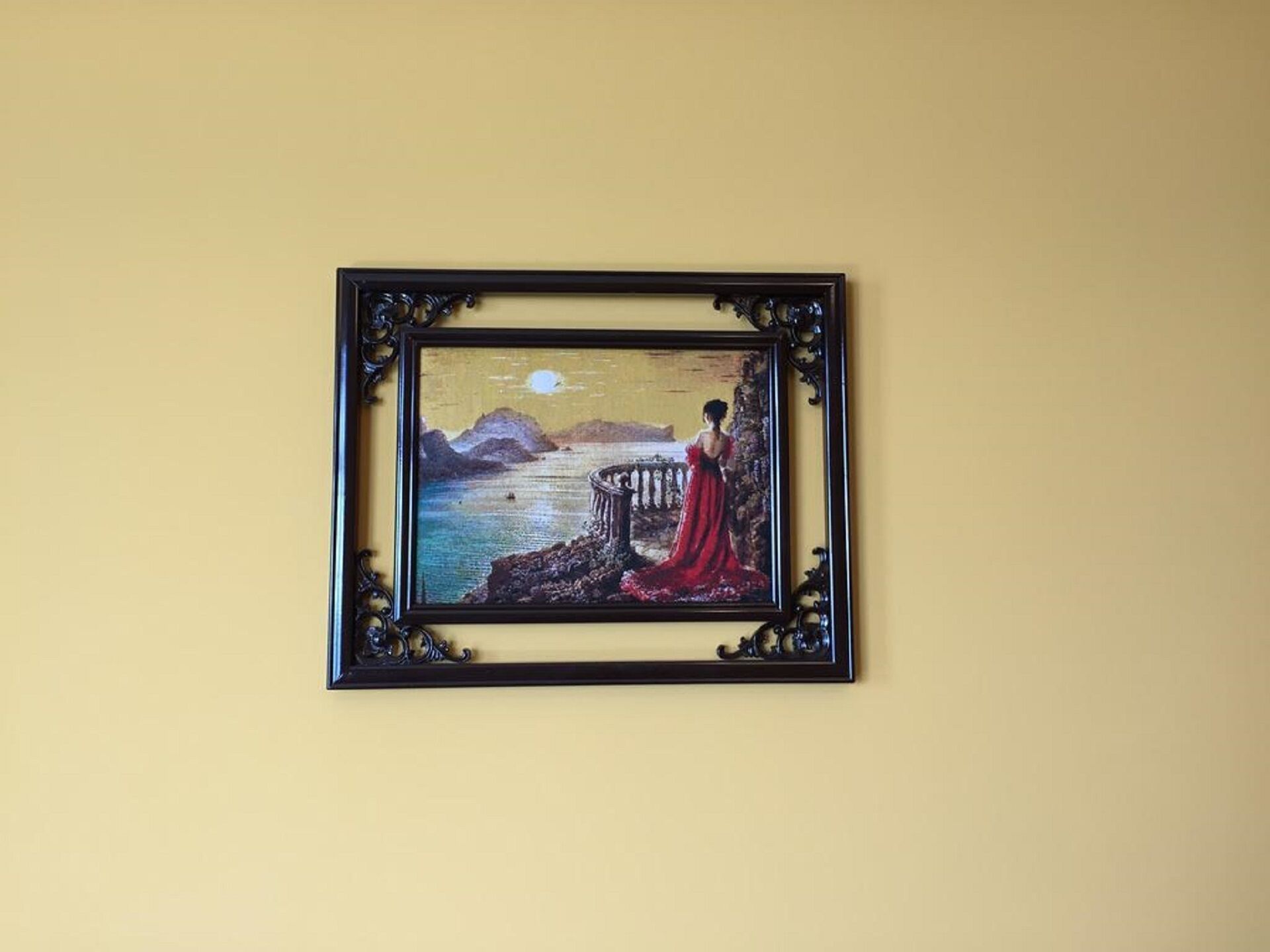 Un tableau encadré représentant une femme vêtue d’une robe rouge, debout près d’une balustrade, d’où elle contemple un plan d’eau avec des montagnes en arrière-plan.