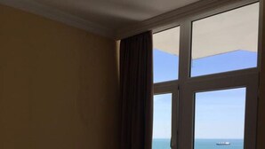 Une chambre d’hôtel avec un lit, une table de chevet, une fenêtre donnant sur la mer et un bateau, et un balcon avec un sol carrelé.