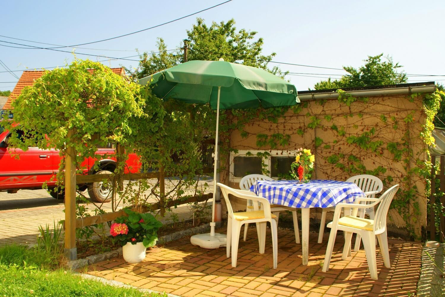 Eine Terrasse im Freien mit Tisch und Stühlen, einem grünen Sonnenschirm und einem in der Nähe geparkten roten Fahrzeug.