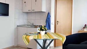 Une cuisine de taille modeste, avec des meubles blancs, une petite table recouverte d’une nappe à rayures, une plante en pot et une serviette bleue accrochée au mur.